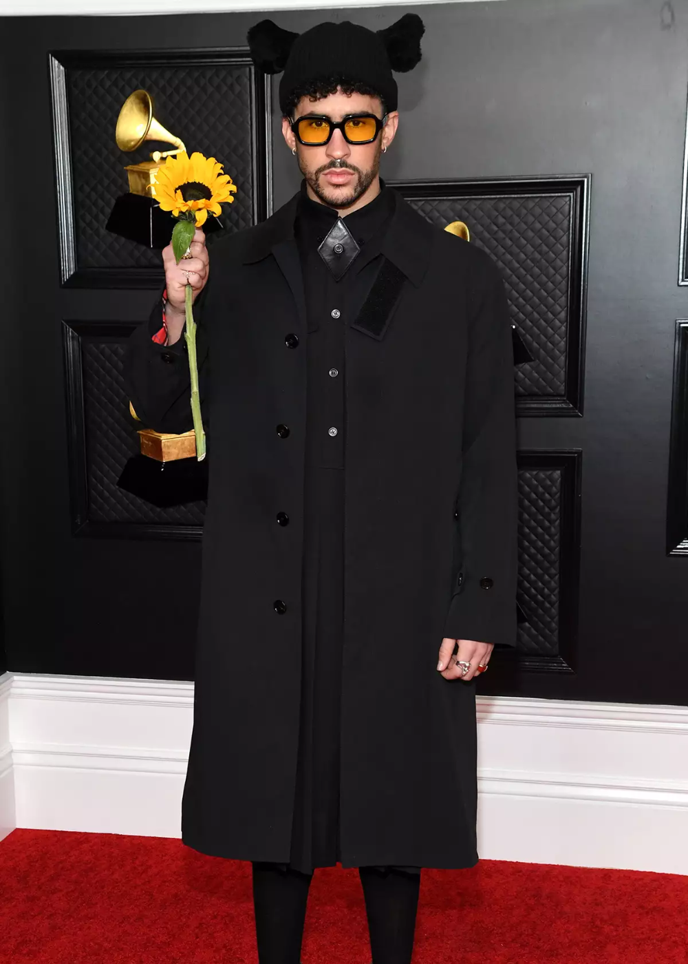 Pessoa com roupa preta segura um girassol no tapete vermelho do Grammy, usando óculos escuros e chapéu preto.