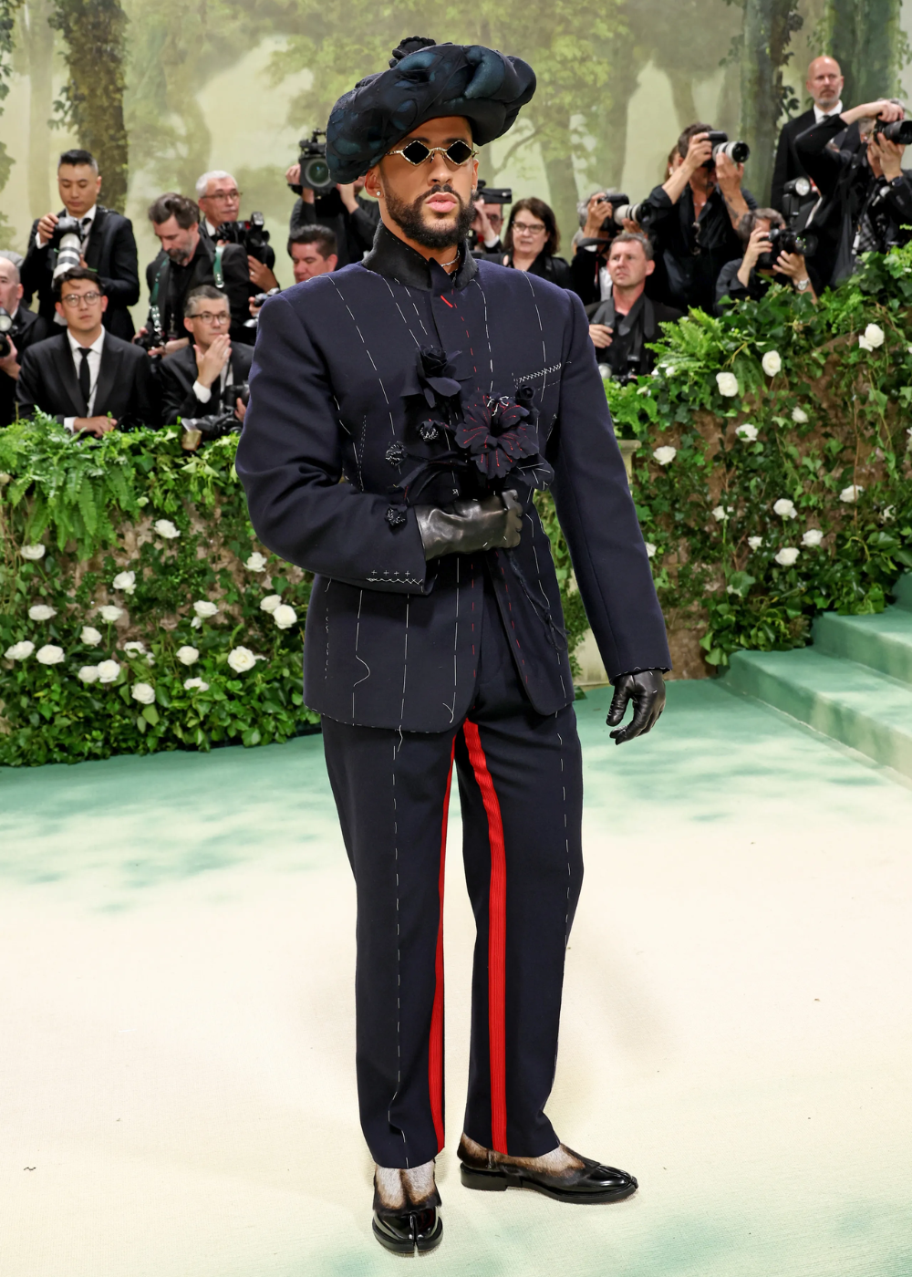 Pessoa com traje sofisticado, chapéu volumoso e óculos escuros em evento elegante, cercada por fotógrafos e vegetação.