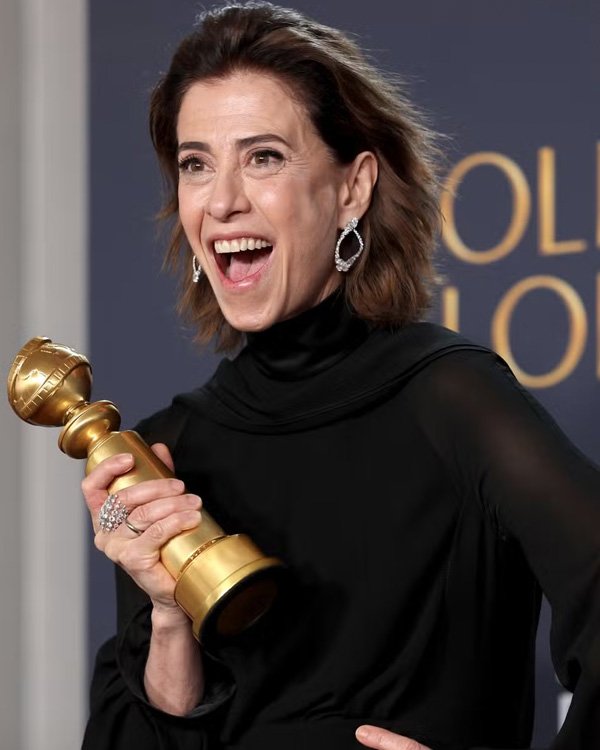 Pessoa sorrindo, segurando troféu dourado com unhas sem esmalte, em traje preto elegante.