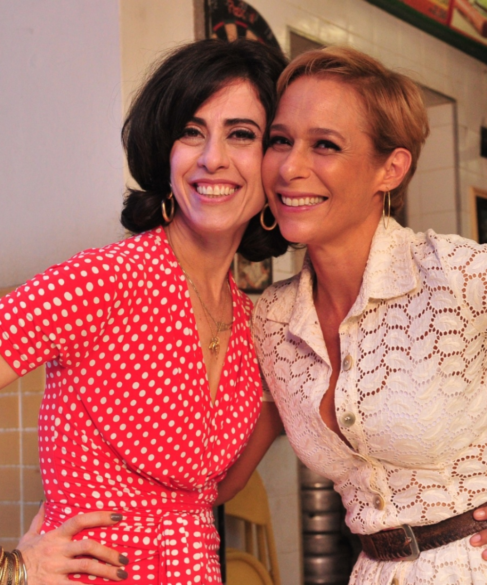 Duas mulheres sorrindo, uma usa vestido de bolinhas e outra vestido claro rendado, abraçadas em local interno.