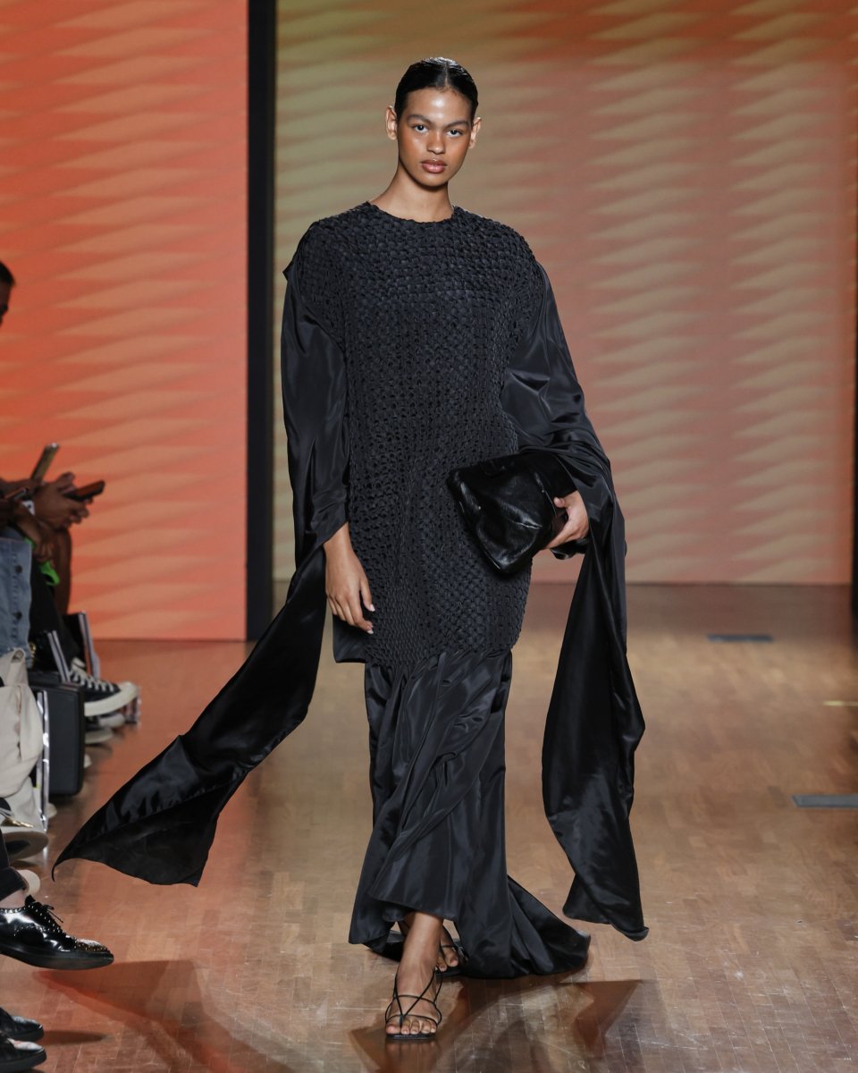 Modelo desfilando em um vestido preto texturizado em uma passarela de madeira clara. Fernanda Torres no Oscar