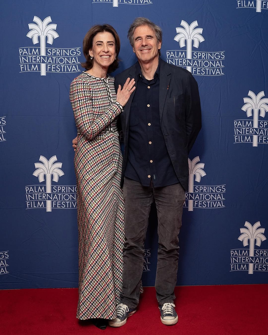 Casal elegante em tapete vermelho do Festival Internacional de Cinema de Palm Springs. Ela veste um vestido estampado. Looks da Fernanda Torres.