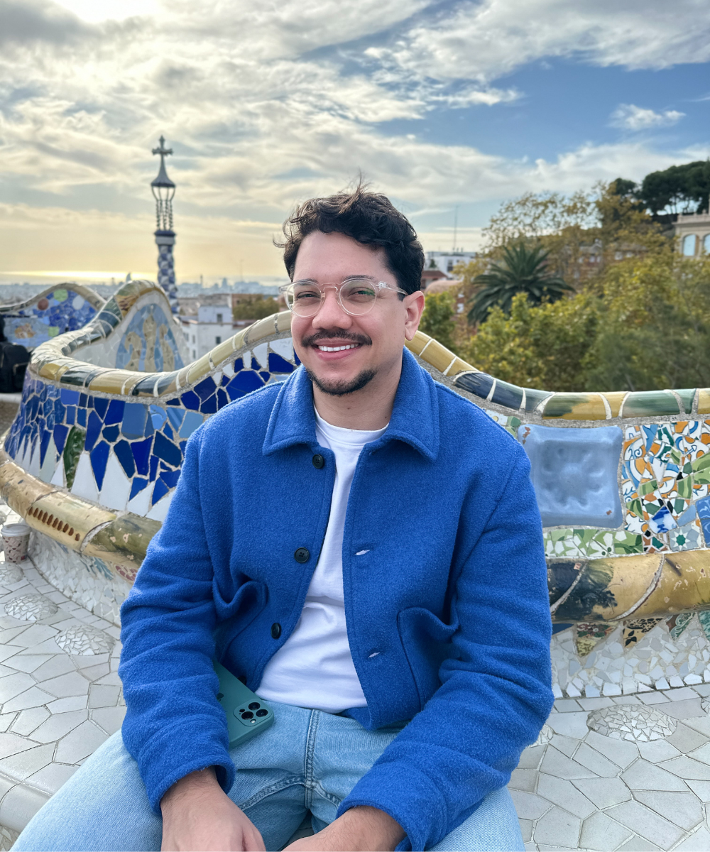 Pessoa sorrindo em um banco colorido no Parque Güell, Barcelona. Fundo com céu azul e natureza.