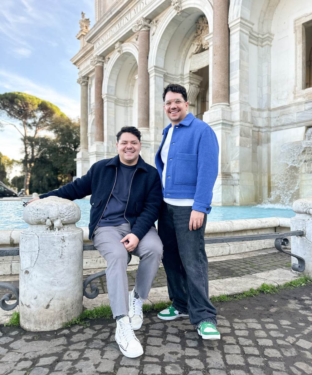 Dois homens sorrindo em frente a uma fonte e arquitetura clássica na Europa, representando uma viagem.