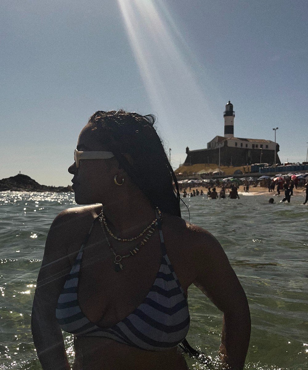 Pessoa em praia de Salvador, com o Farol da Barra ao fundo, aproveitando o sol e o mar.