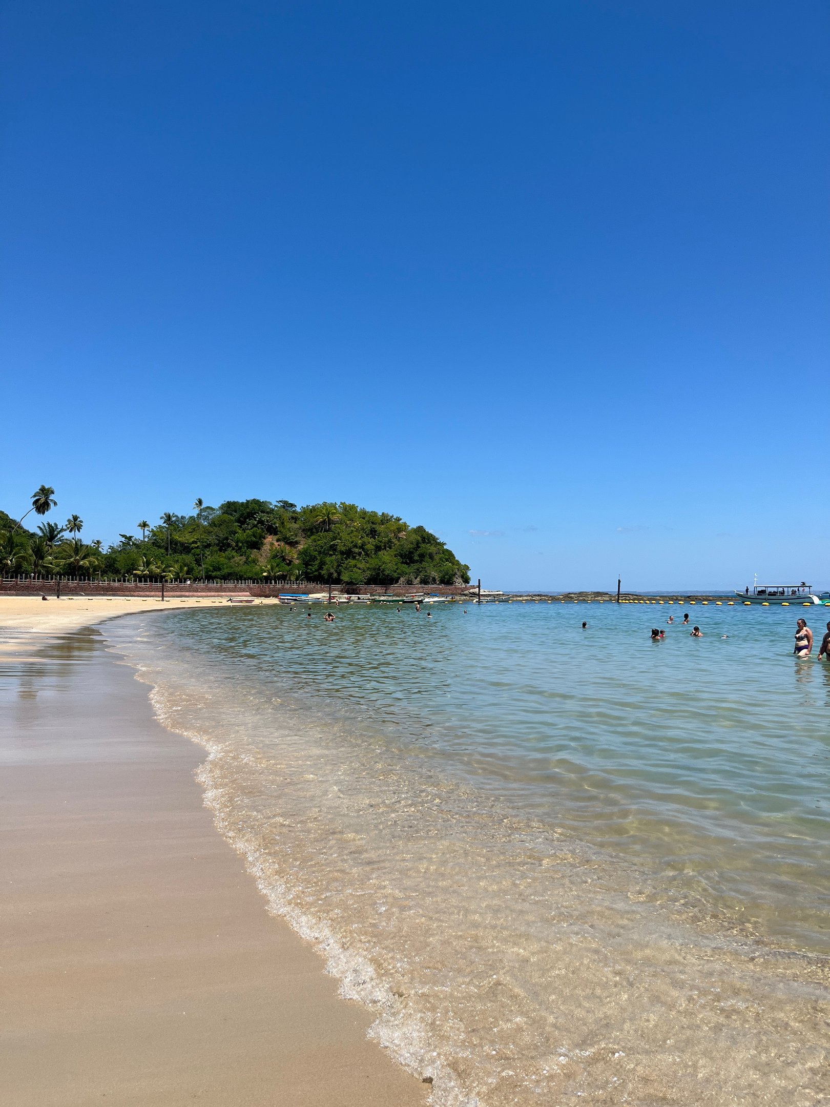 Praia com areia clara e águas tranquilas, pessoas nadando, céu azul. Atividade popular em Salvador.