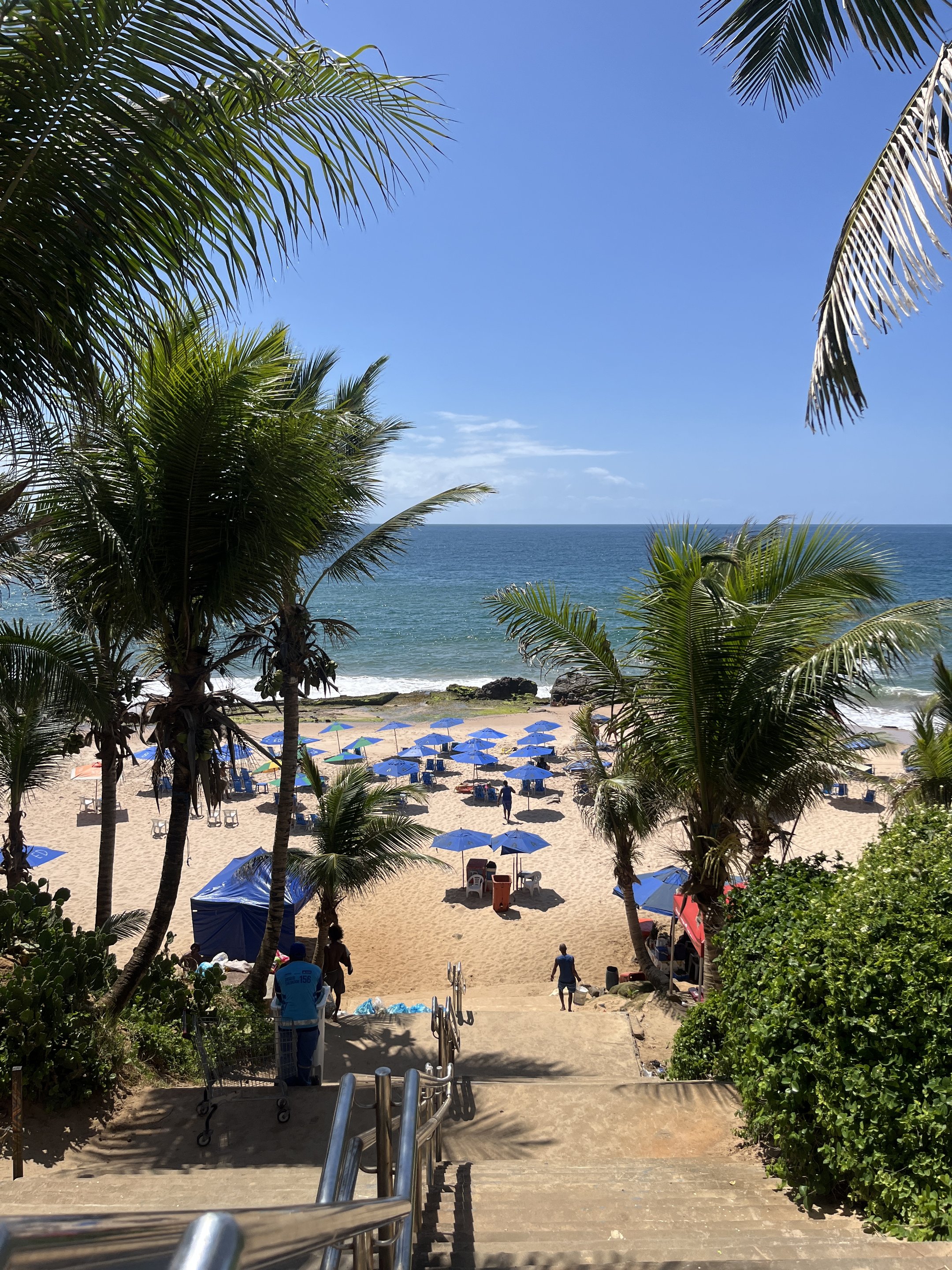 Praia com palmeiras, guarda-sóis azuis e mar azul, em Salvador.