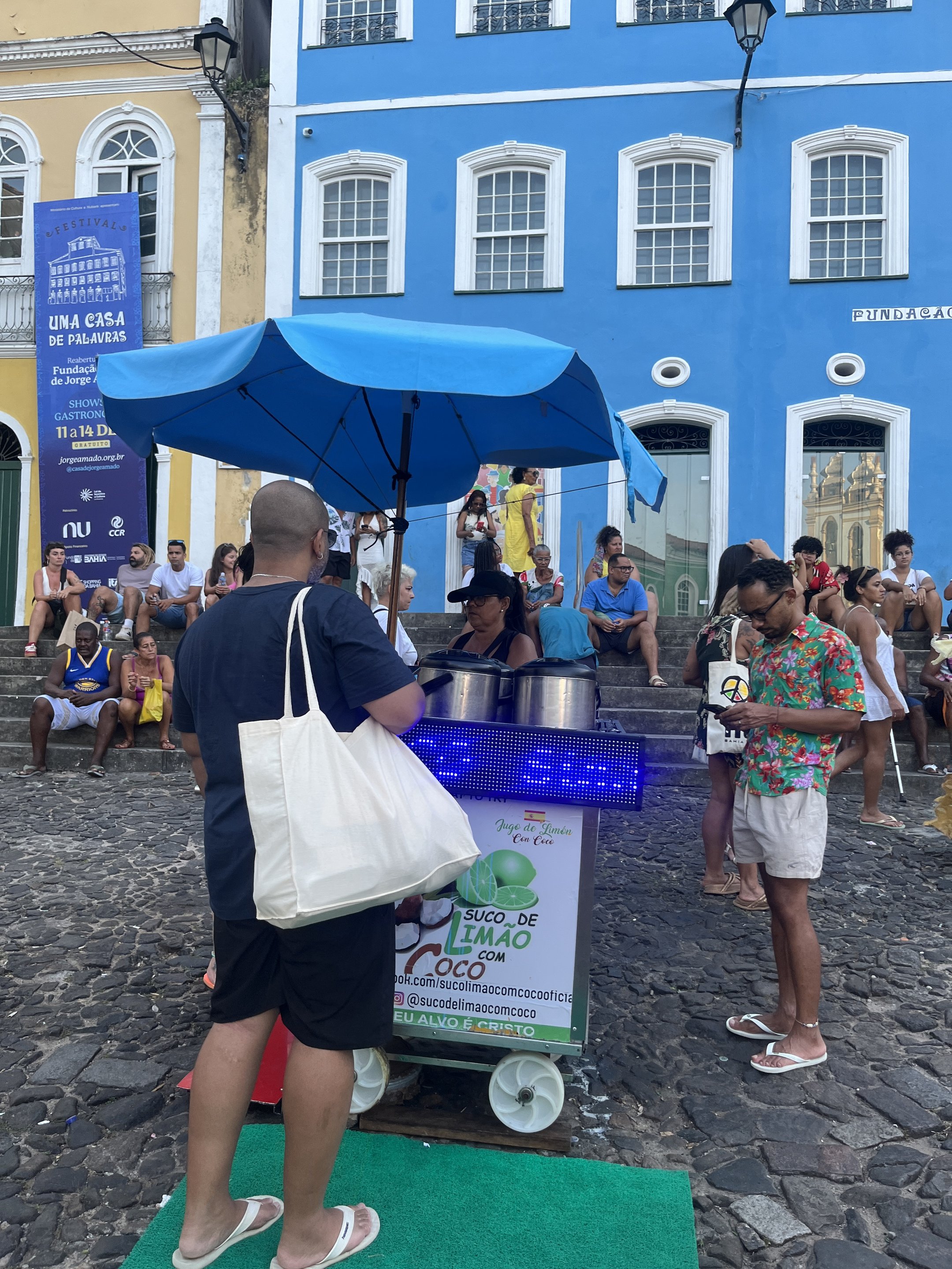 Carrinho de suco em rua de Salvador, com pessoas reunidas ao redor e prédios coloridos ao fundo.