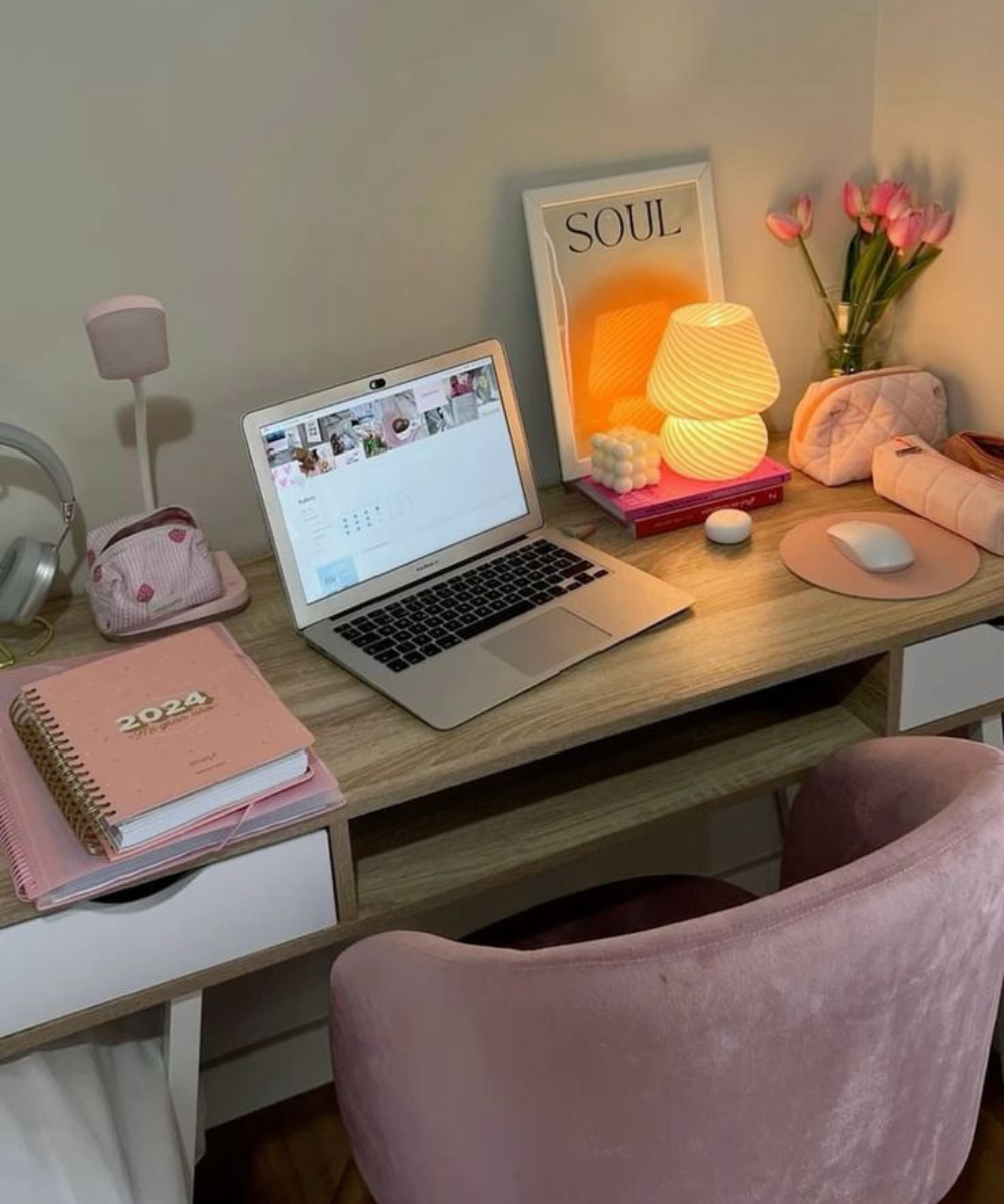 Mesa de trabalho com laptop, abajur aceso, flores, e itens de papelaria como agenda rosa e estojo ao lado.