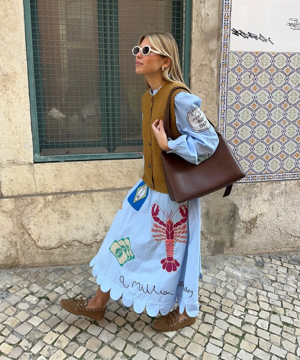 Vestido azul com estampa de lagosta vermelha, complementado por colete marrom, bolsa e óculos escuros.