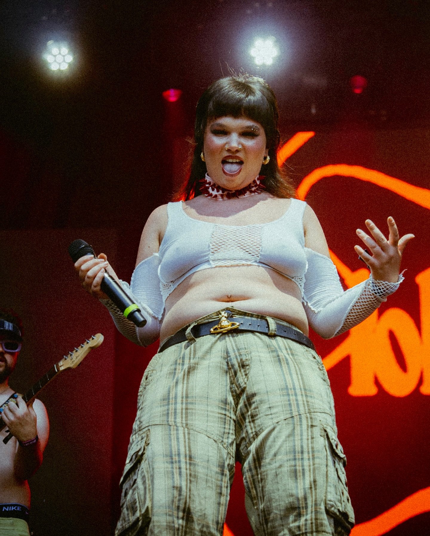 Cantora performa em palco, usando top branco e calça xadrez. Luzes e instrumentos ao fundo criam atmosfera vibrante.