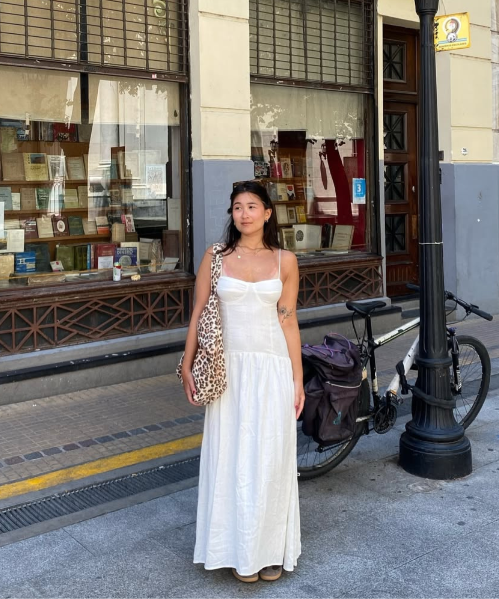 Mulher de vestido branco em rua ensolarada, perto de bicicleta e vitrine de livros, destacando o clima de viajar no verão.