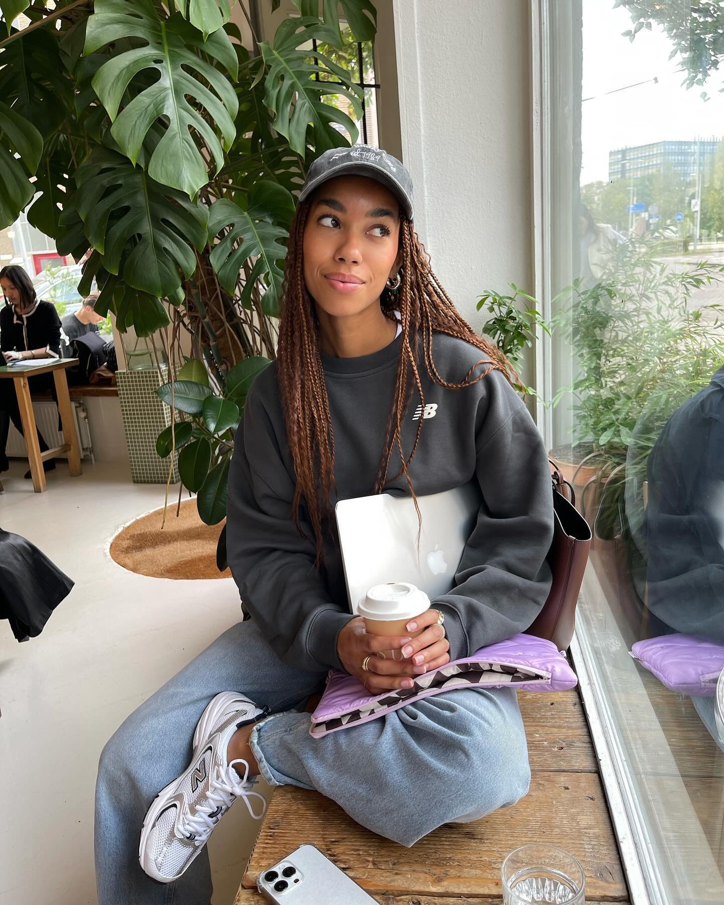 Mulher sentada em café, com tênis estilo anos 90, casaco cinza, boné e laptop; segurando copo de café.