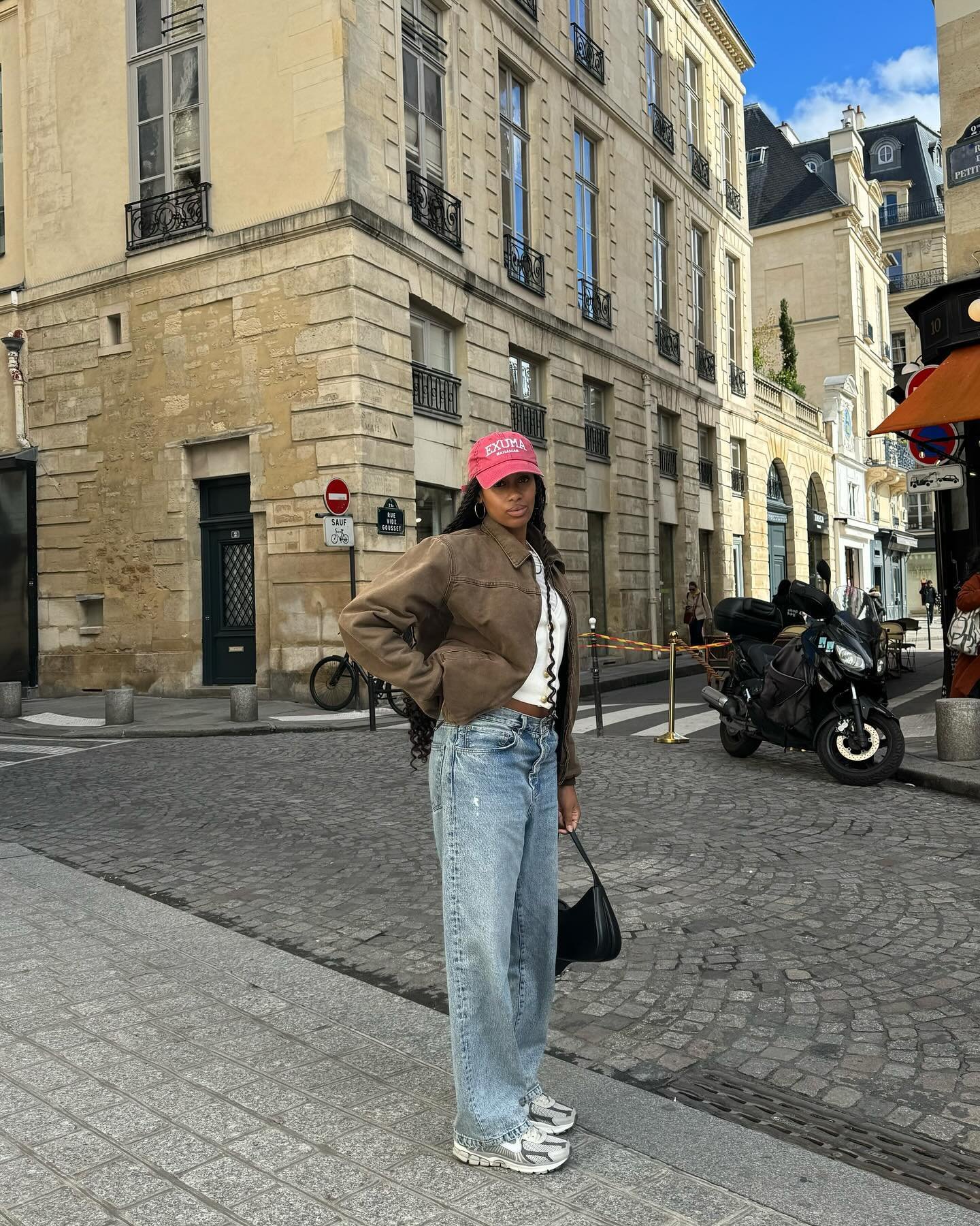 Pessoa com boné vermelho e tênis estilo anos 90, em uma rua de paralelepípedos com prédios históricos.