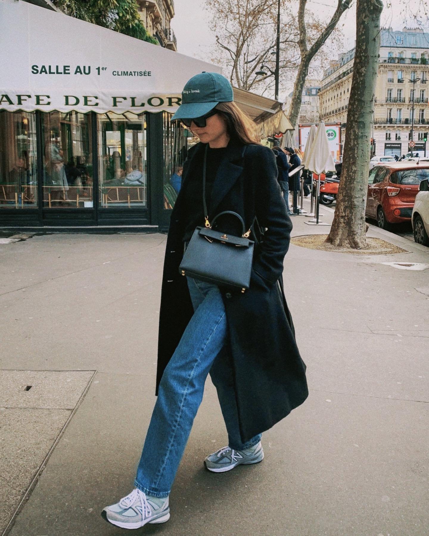 Mulher caminhando em Paris com jeans, casaco longo e boné, usando tênis estilo dos anos 90.