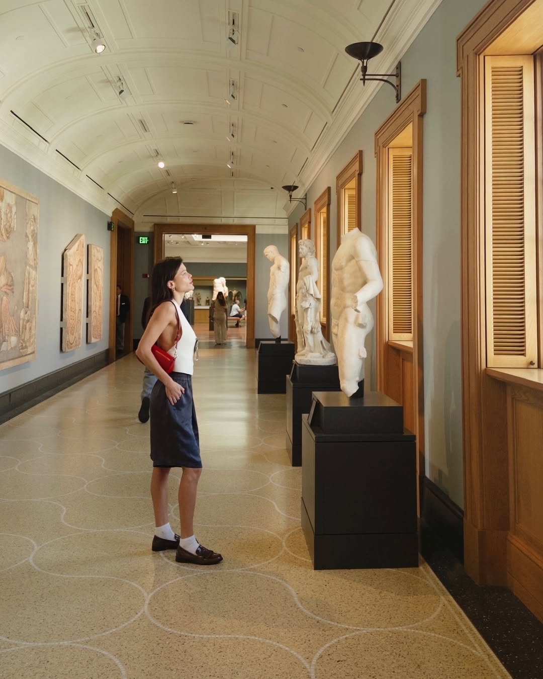 Pessoa observa esculturas em um corredor de museu, com teto abobadado e luz natural entrando pelas janelas. Vitoria Strada BBB25