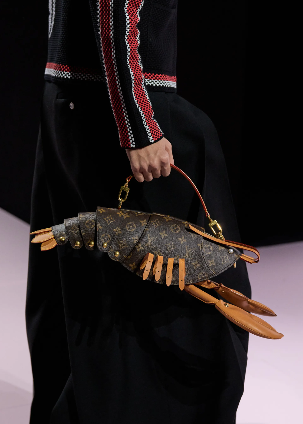 Bolsa em formato de lagosta com estampa monograma é carregada por uma pessoa vestindo jaqueta de detalhes vermelhos.