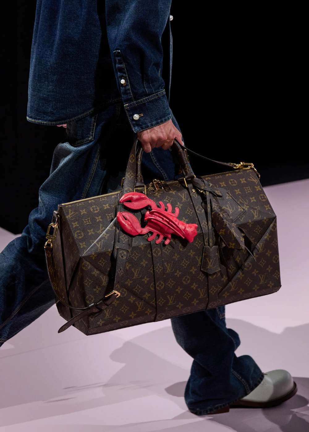 Bolsa marrom estampada sendo carregada por pessoa usando jeans, com adorno de lagosta vermelha presa.