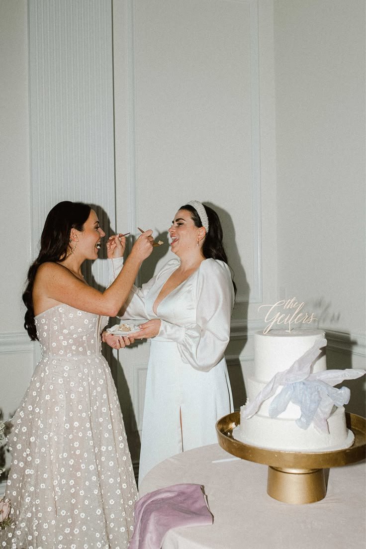 Duas mulheres sorridentes trocam pedaços de bolo em um casamento, ao lado de um bolo decorado com flores. Melhor lua para casar.