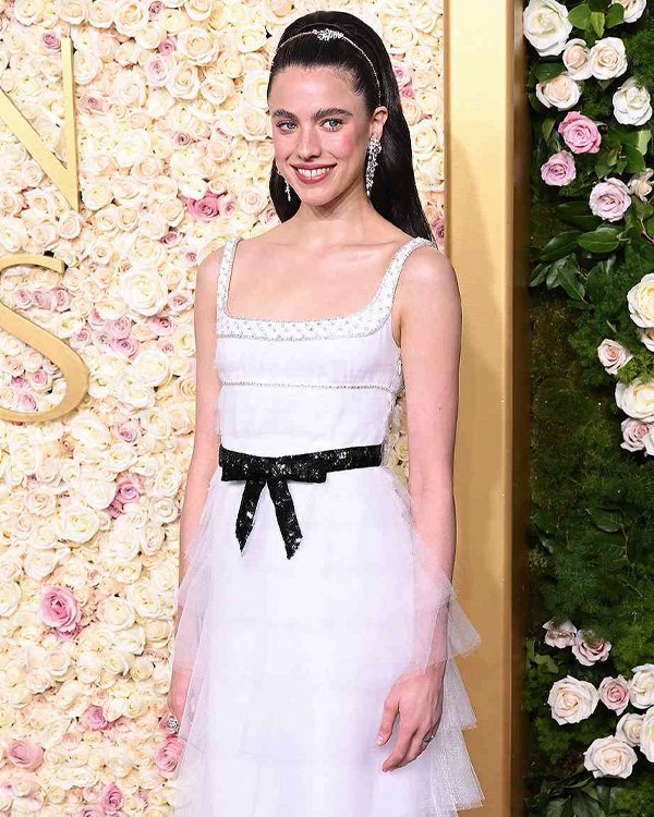 Uma mulher sorrindo em frente a um fundo florido, usando vestido branco com cinto preto. Unhas sem esmalte.