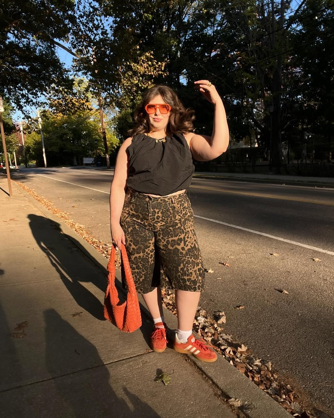 Mulher com shorts de oncinha, top preto, óculos laranja e maxi bolsa vermelha andando na calçada ensolarada.