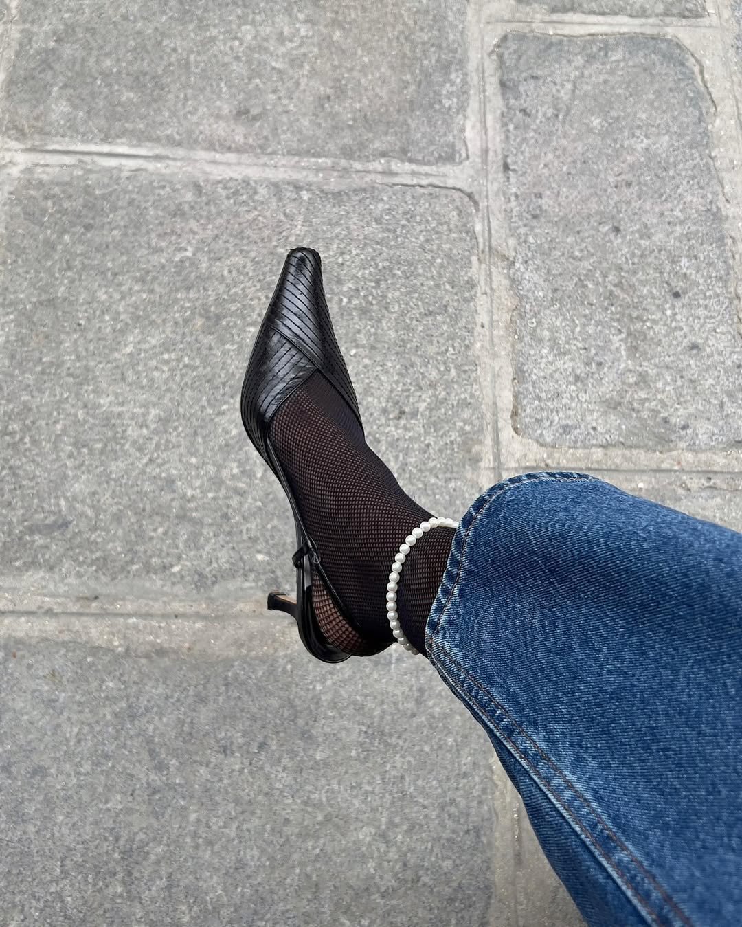 Sapato preto com meia arrastão e calça jeans em piso de pedra, destacando elegância e simplicidade dentro das tendências minimalistas.