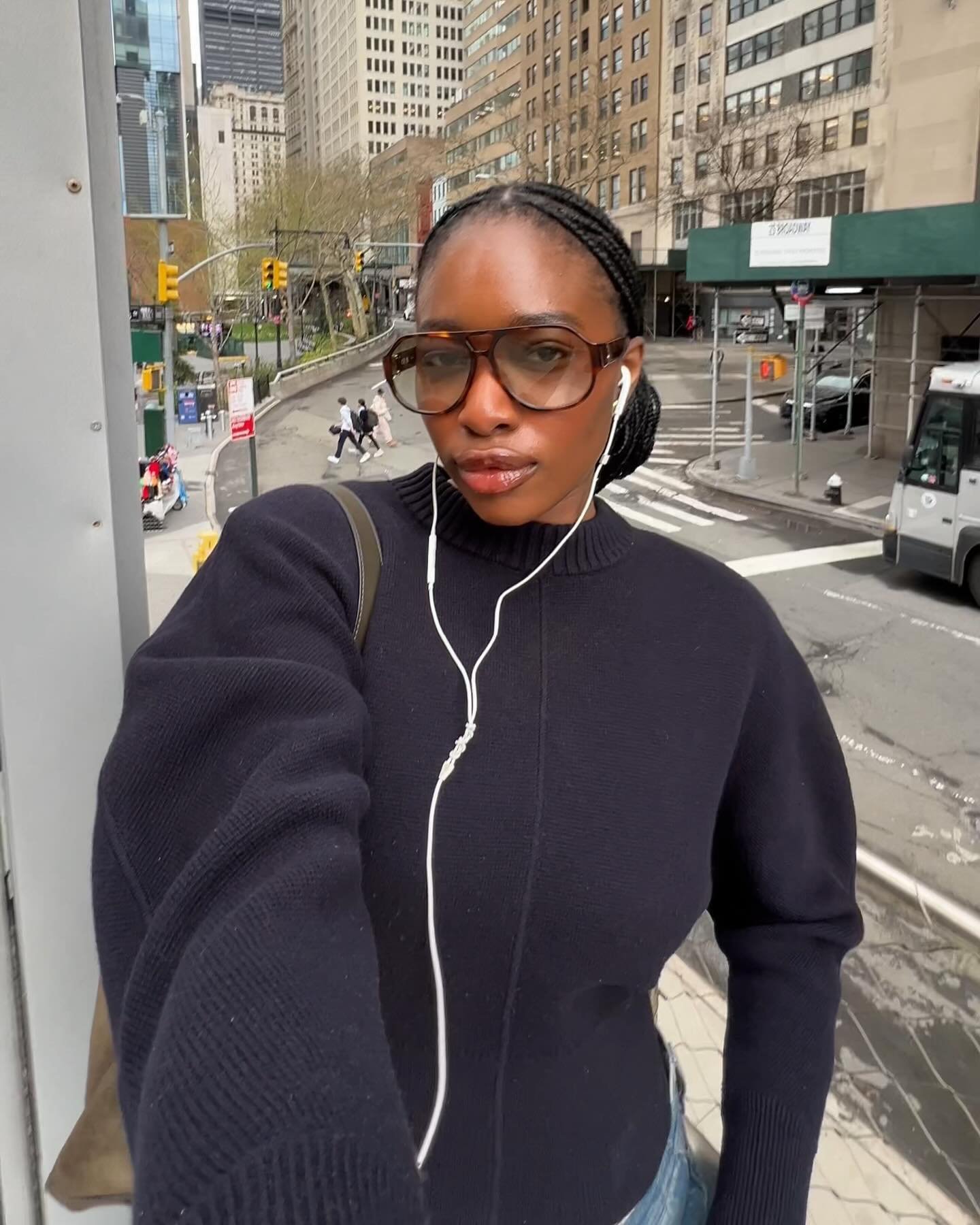 Mulher usando óculos clássicos e fones de ouvido, caminhando em uma rua movimentada da cidade.