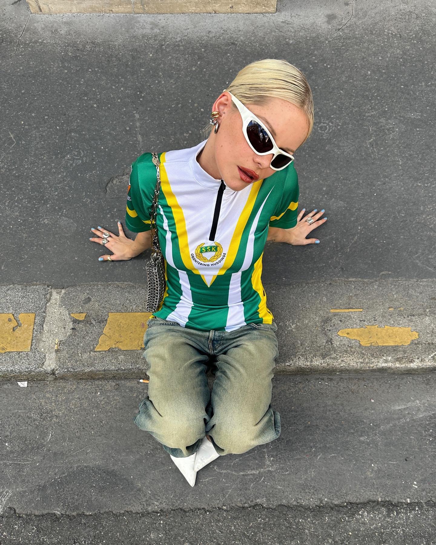 Pessoa com cabelo loiro e óculos clássicos brancos usa camisa verde e amarela sentada no meio-fio.