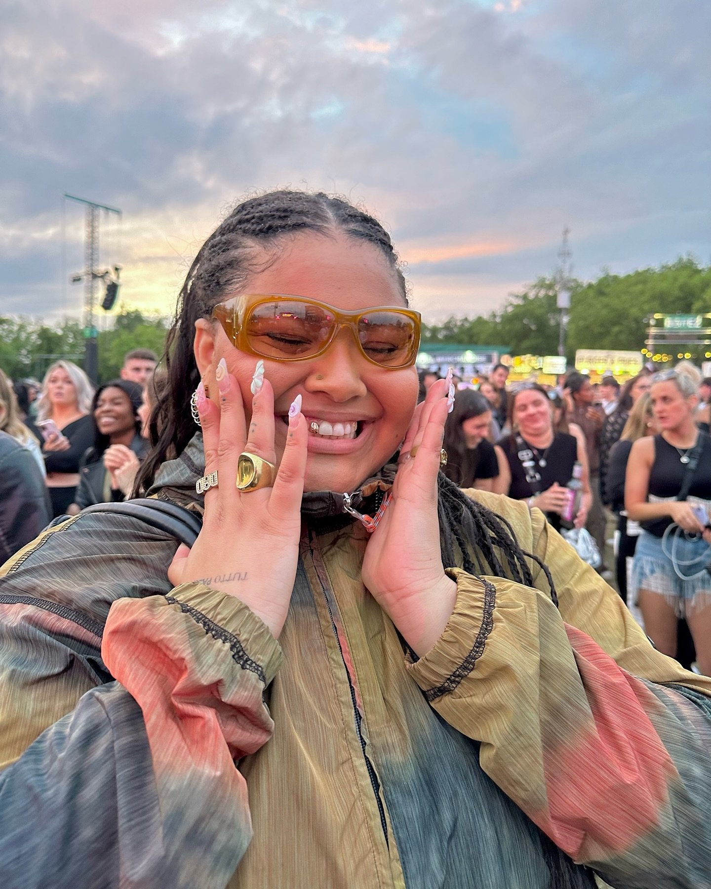 Pessoa sorridente com óculos de sol, cercada por uma multidão ao ar livre, aparenta estar em um evento festivo.