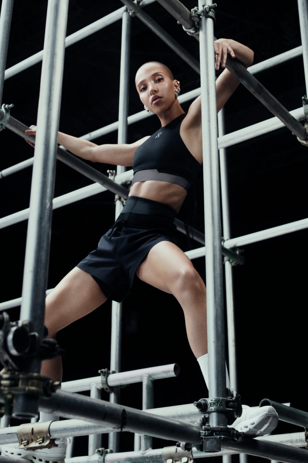 Pessoa em pose dinâmica entre estruturas metálicas, usando roupa esportiva preta. Estilo da colaboração On x Fka Twigs.