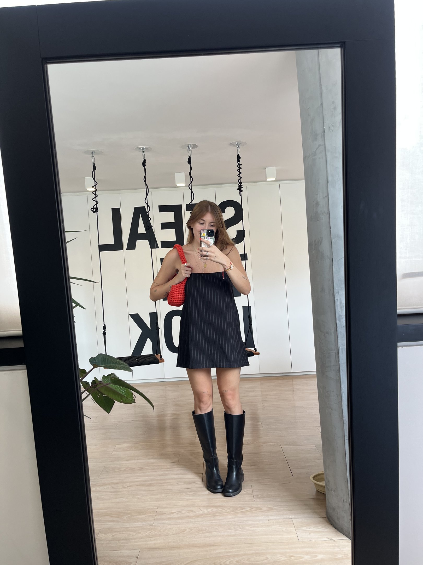 Pessoa vestindo vestido preto e botas longas, segurando bolsa vermelha, se olhando no espelho.