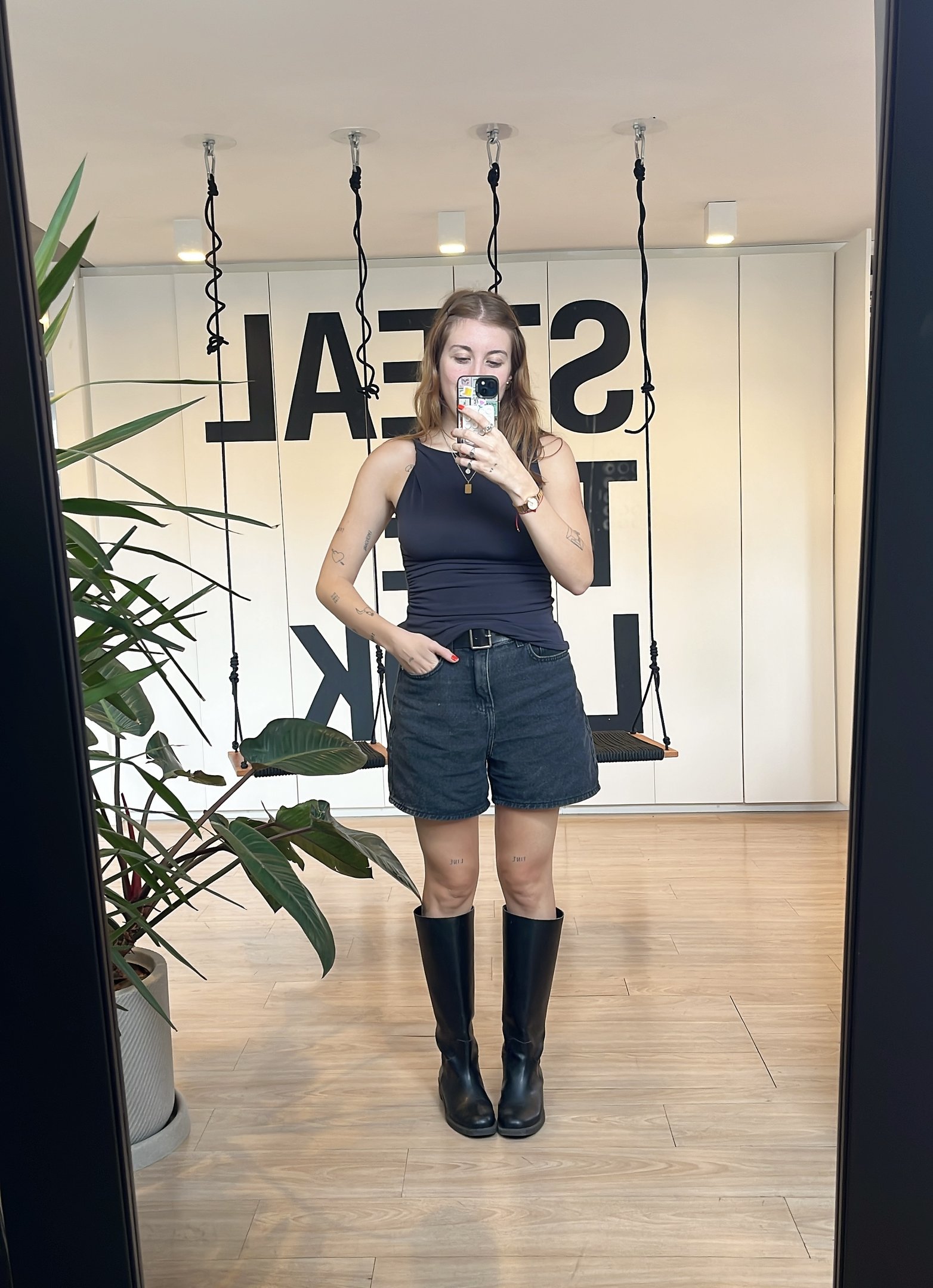 Pessoa vestindo regata preta, shorts jeans e botas pretas em frente a um espelho com plantas ao lado.
