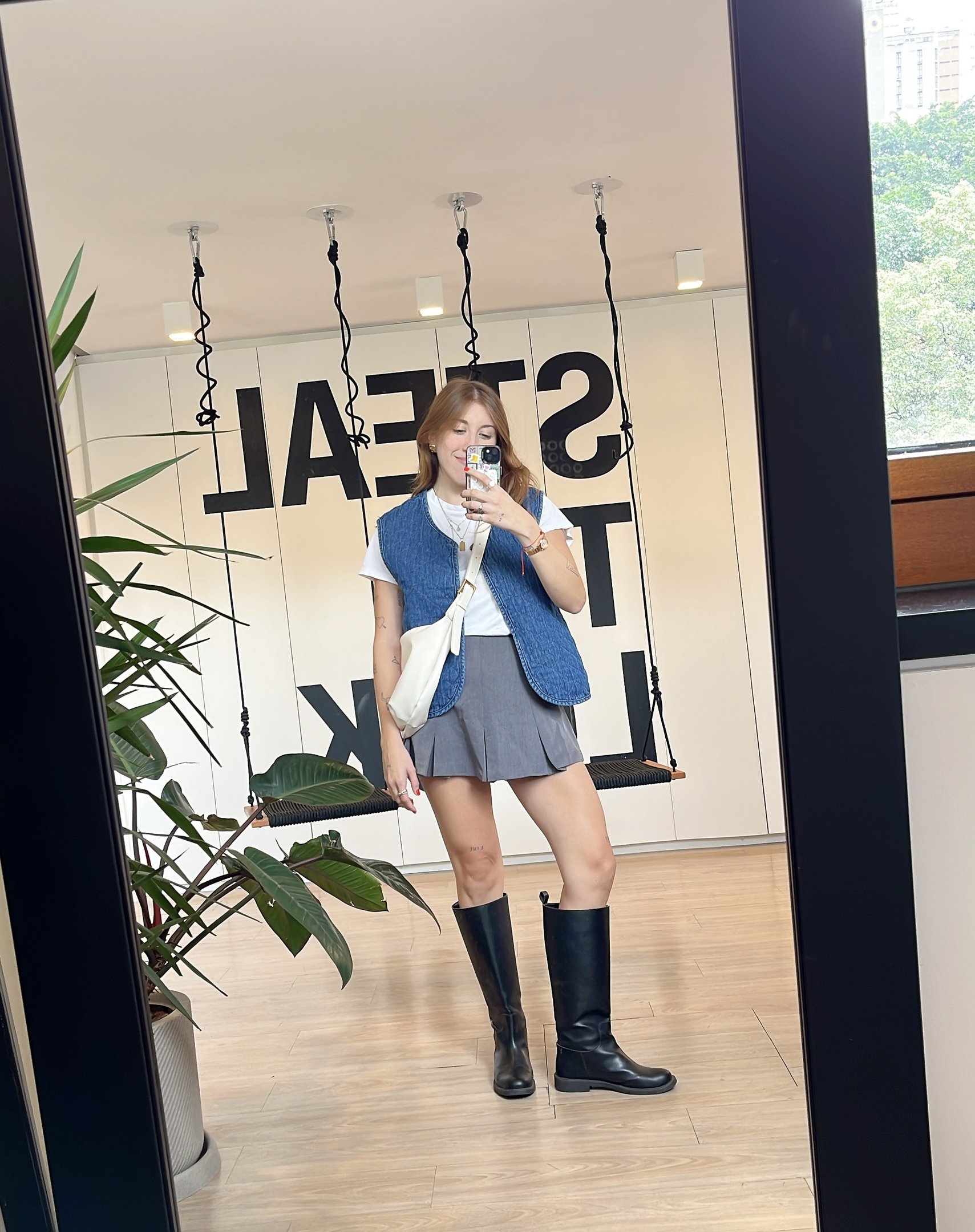 Pessoa usando looks com bota e colete azul, saia curta cinza. Ela tira foto em frente ao espelho com balanços ao fundo.