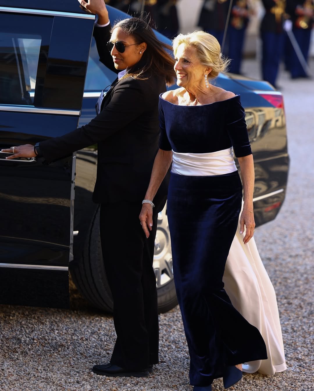 primeiras-damas em traje formal, uma abrindo a porta do carro e outra sorrindo em um vestido azul e branco, ambiente ao ar livre.