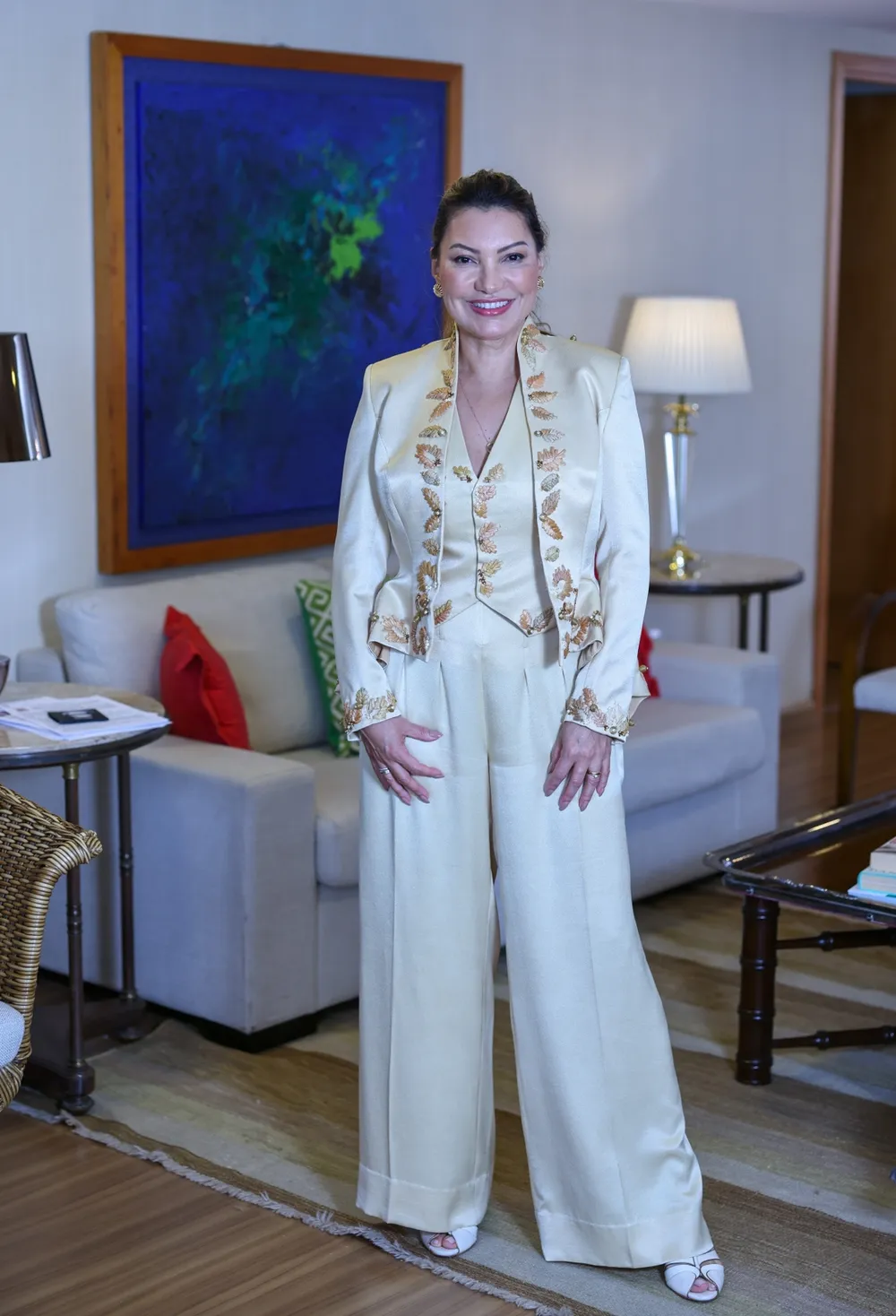 Mulher em traje formal decorado com bordados dourados, em uma sala de estar elegante.