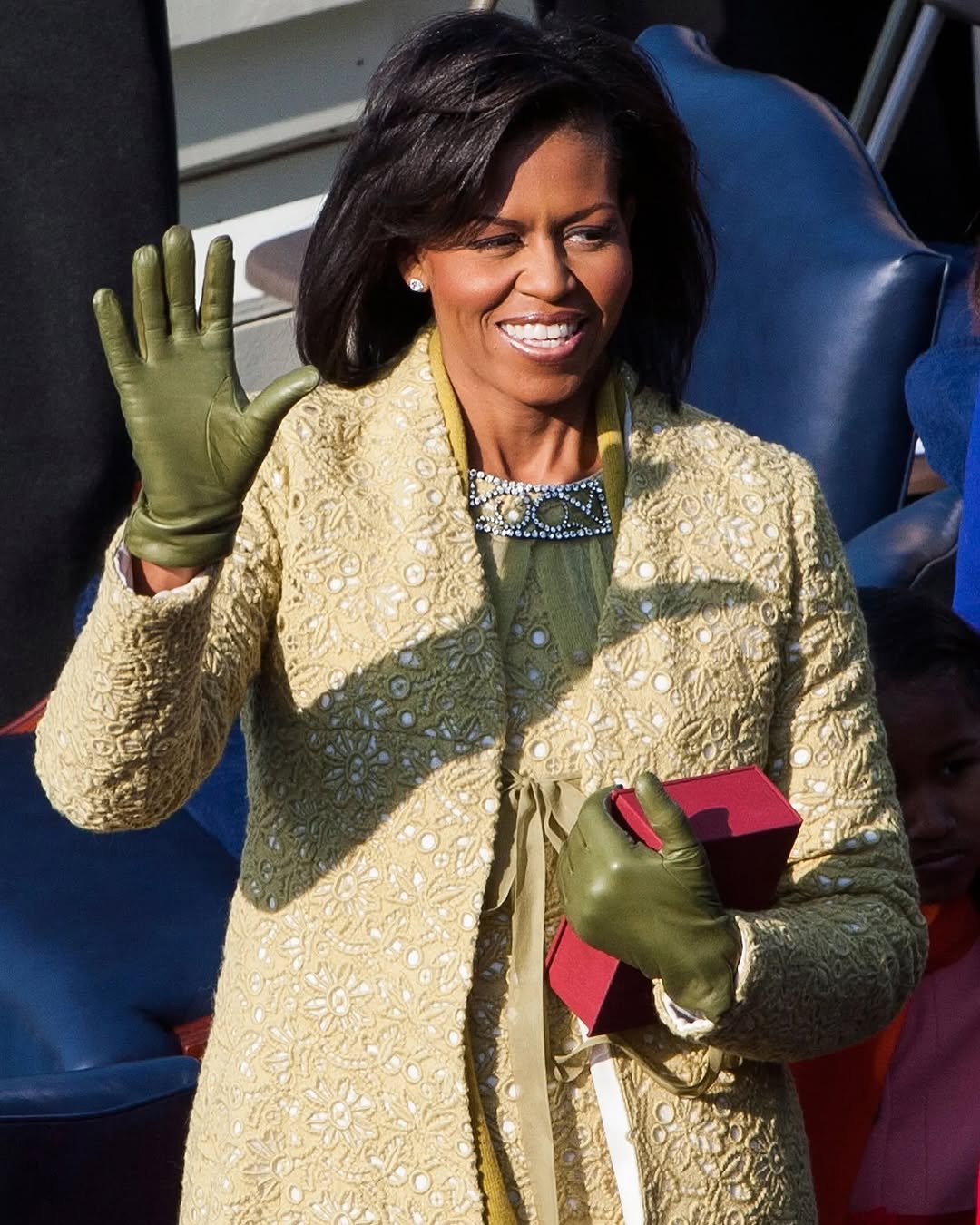 Michelle Obama, uma das primeiras-damas sorridente usando casaco elegante e luvas verdes, levantando a mão.