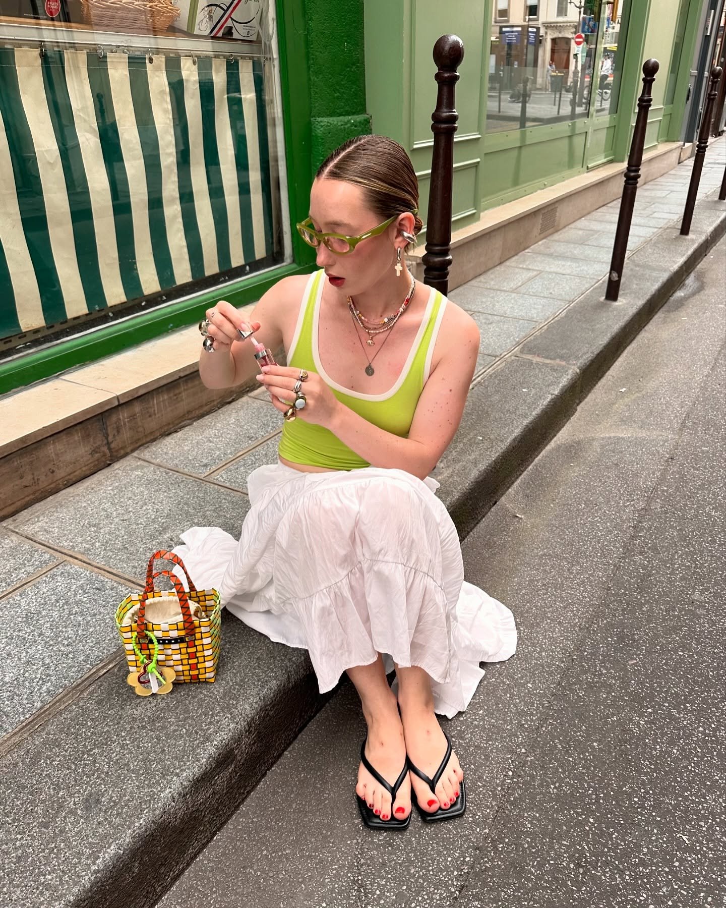 Pessoa sentada em calçada usando saia branca, regata verde-limão e chinelos. Bolsa colorida ao lado, transmitindo roupas estilosas e confortáveis.