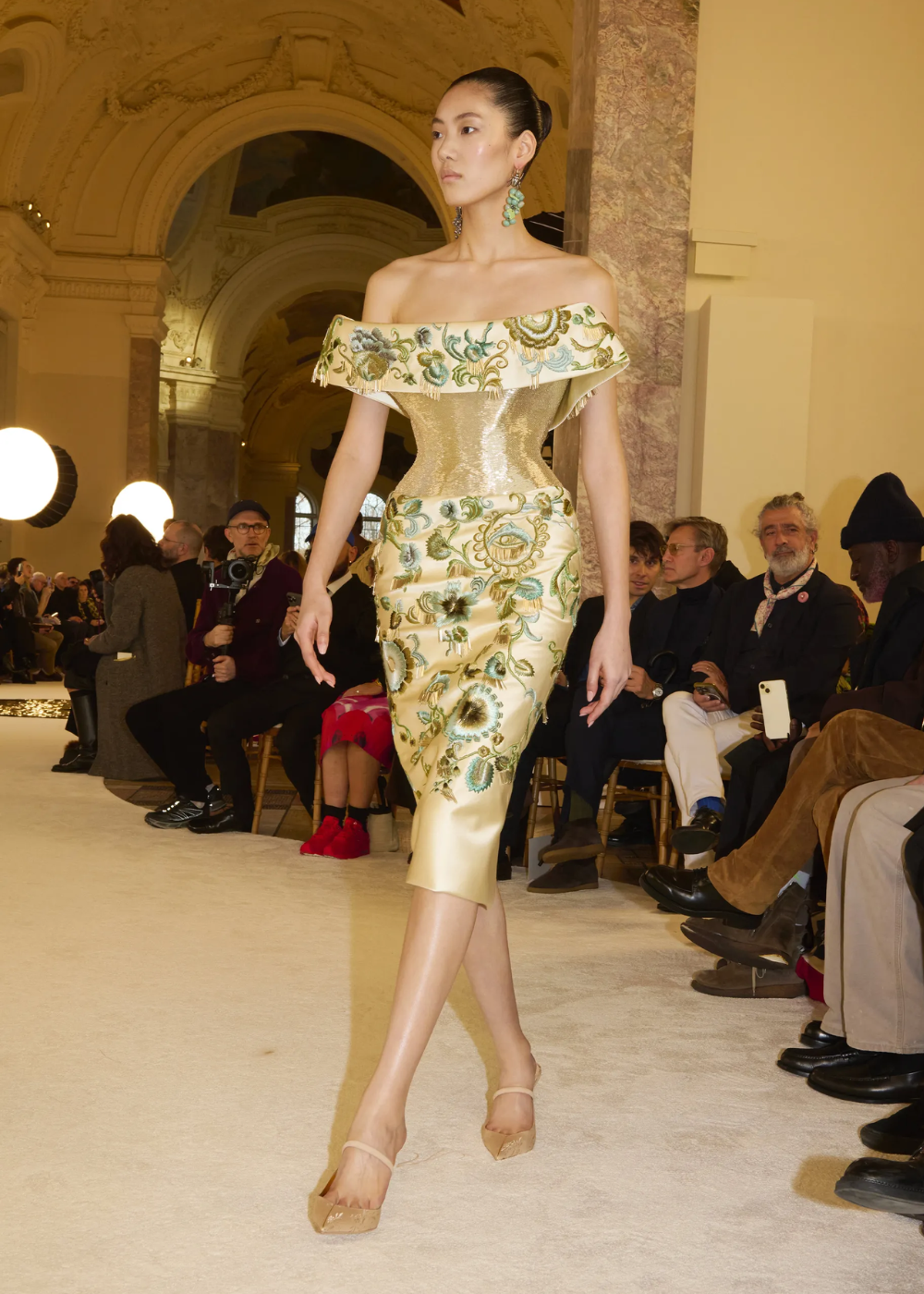 Modelo em vestido dourado com bordados florais, desfilando em evento da Semana de Alta-Costura 2025.