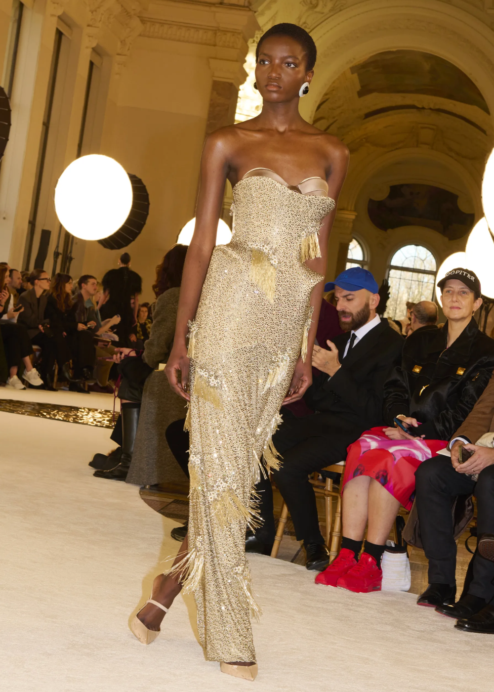 Modelo desfila em vestido dourado com franjas brilhantes em evento de moda, inspirado na Semana de Alta-Costura 2025.