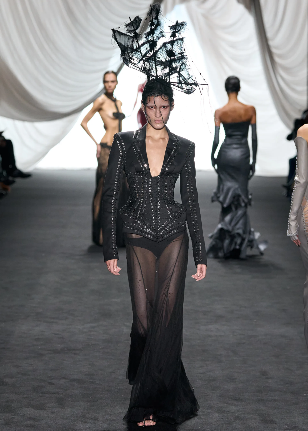 Modelo veste visual preto elegante com detalhes metálicos, incluindo chapéu artístico. Evento da Semana de Alta-Costura 2025.