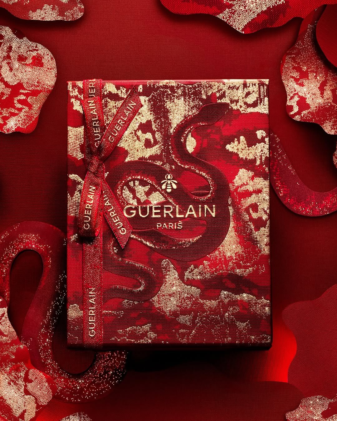 Caixa decorativa Guerlain em tons de vermelho e dourado, com padrões ondulantes e laço vermelho impresso com o nome da marca.