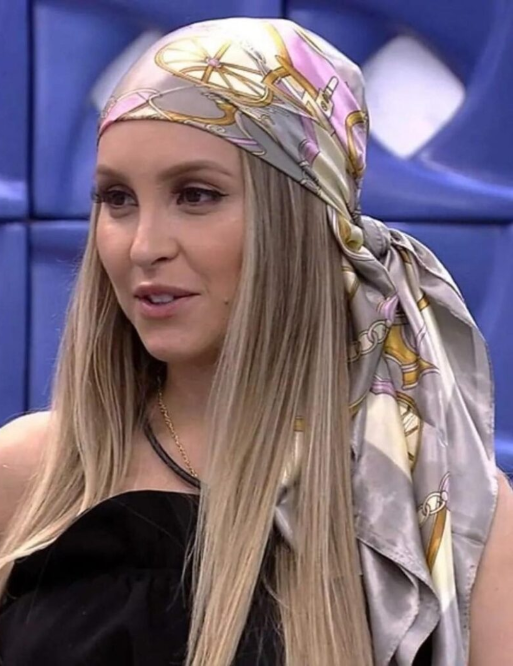 Lenço colorido na cabeça, estilo marcante do BBB, com tendência de moda destaque. Cabelo loiro liso e blusa preta.