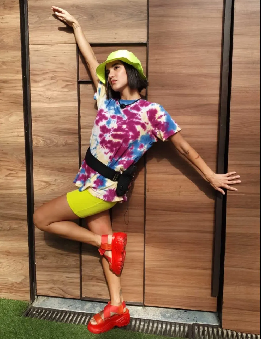 Mulher com blusa tie-dye, shorts ciclista amarelo, chapéu verde e sandálias vermelhas. Visual lembra tendências de moda que marcaram o BBB.