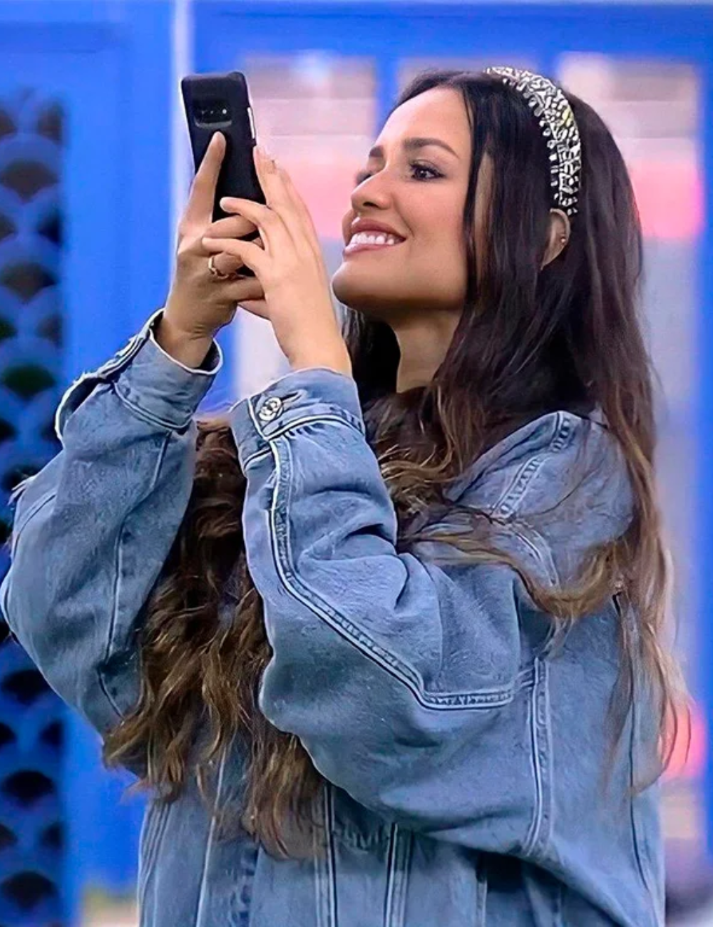 Mulher sorridente, vestida com jaqueta jeans e tiara brilhante, usa celular. Combinação casual e estilosa.