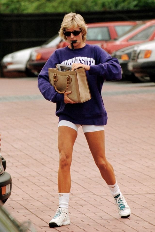 Pessoa andando na rua com moletom roxo e shorts branco, segurando bolsa grande. Calça tênis dos anos 90.