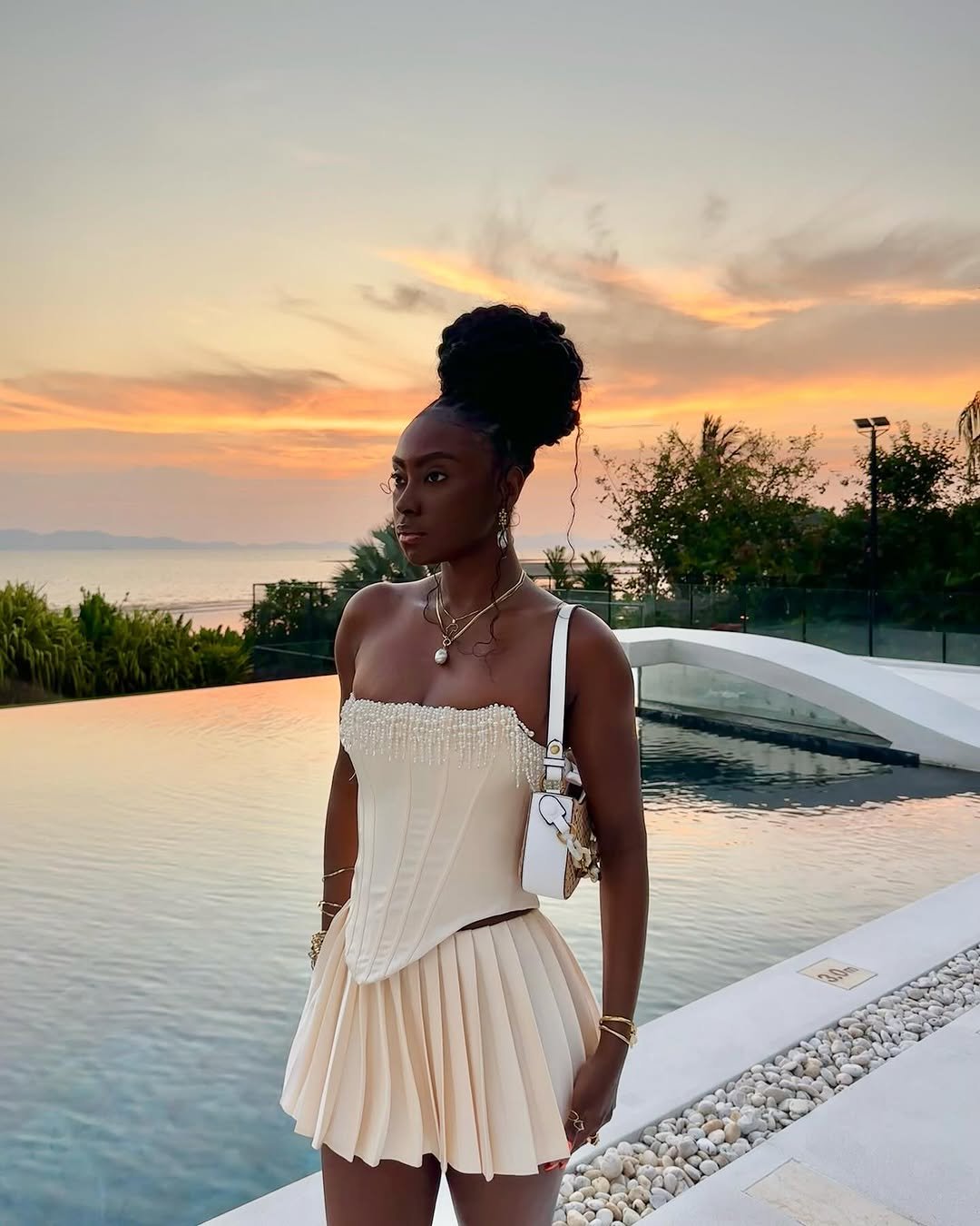 Mulher com cabelo preso, em frente à piscina e pôr do sol ao fundo. Trança na praia