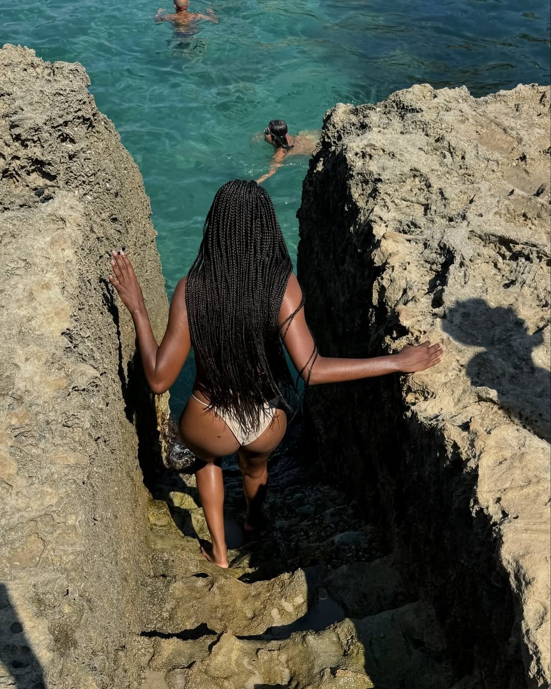 Pessoa com Trança na praia desce degraus entre rochas em direção ao mar cristalino, na praia.