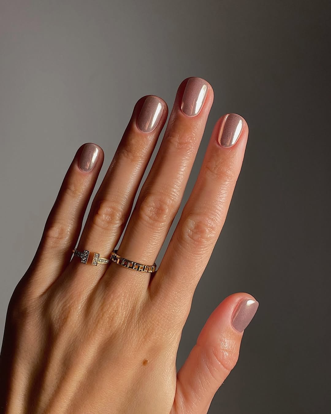 Mão com unhas básicas esmaltadas em tom nude, usando anéis dourados.