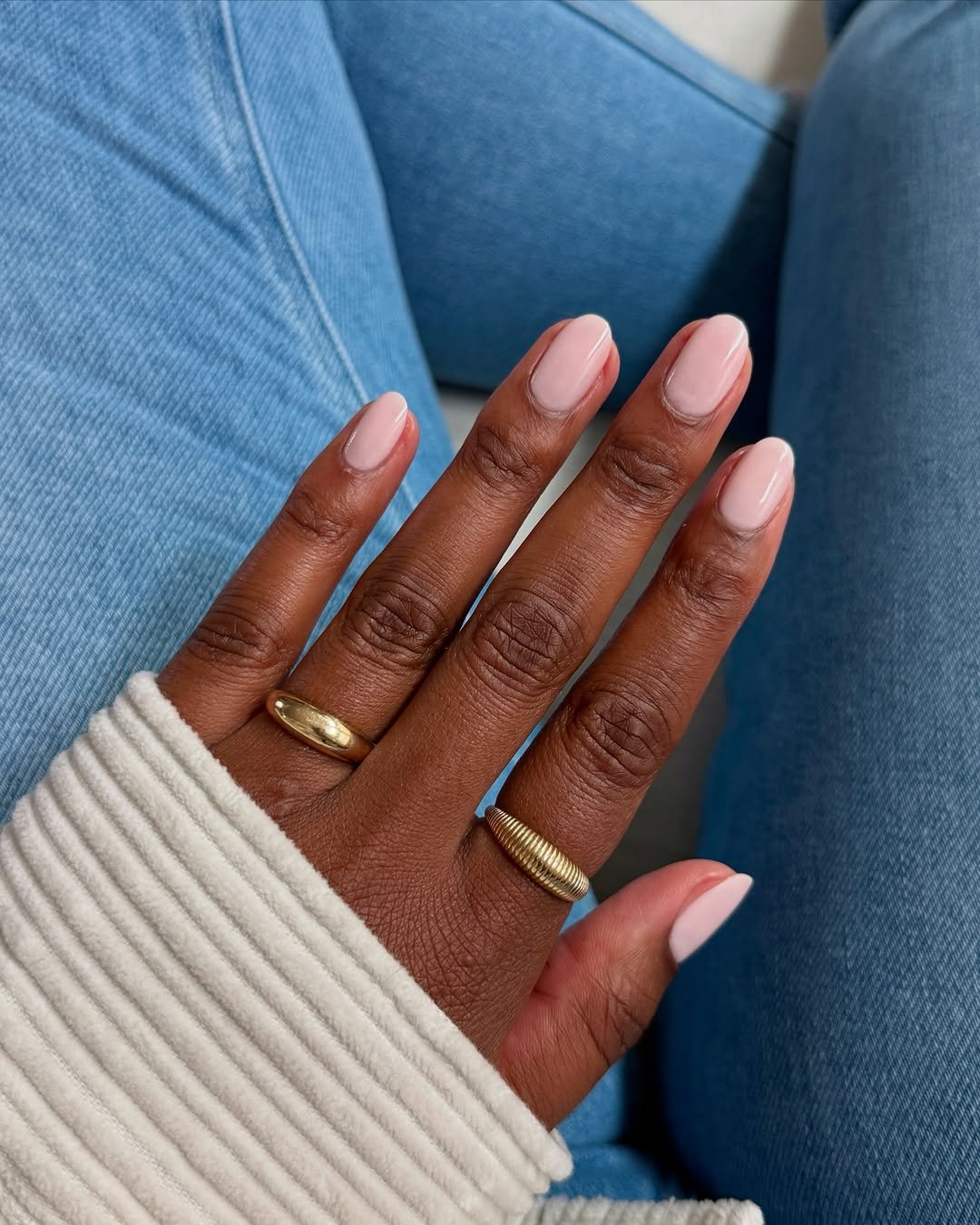 Mão com unhas básicas em esmalte claro, usando anéis dourados, sobre calça jeans azul.