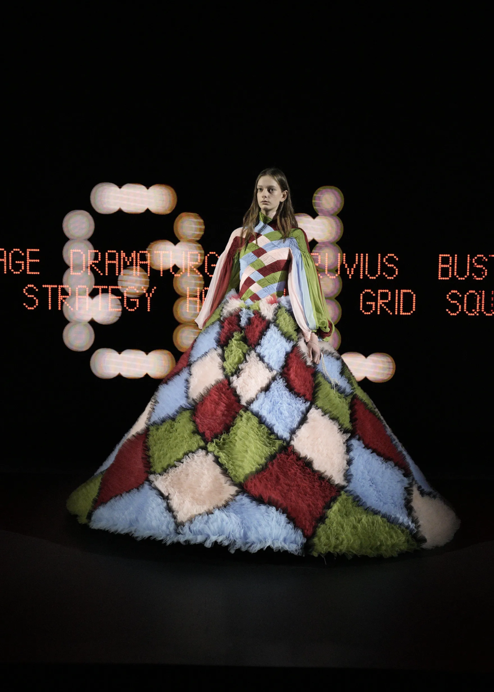 Modelo em vestido colorido com padrão geométrico em desfile da Semana de Alta-Costura 2025. Texto digital iluminado ao fundo.